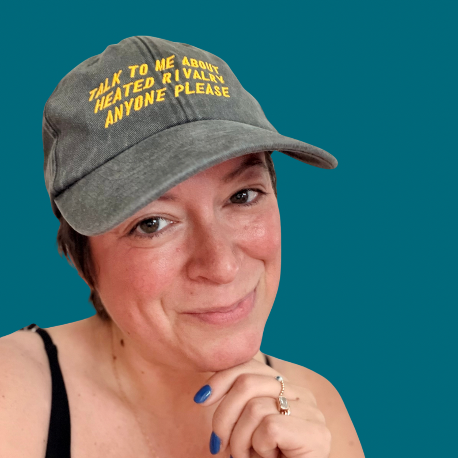 A woman wearing a gray baseball cap with yellow embroidered text that reads 'TALK TO ME ABOUT HEATED RIVALRY ANYONE PLEASE' against a solid teal background.